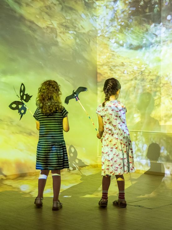 Photo: Back view of two children standing in front of a play of light and shadow in yellow-green colours with two butterflies projected onto the wall.