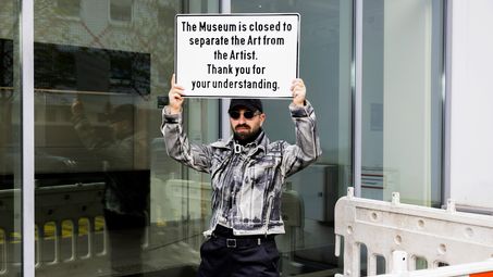 Cem A., "Thank you for your understanding", 2023 A person stands in front of a glass façade holding a sign above their head that reads “The Museum is closed to separate the Art from the Artist. Thank you for your understanding.” In the foreground, there are barriers; the person wears sunglasses, a cap, and a gray patterned jacket.