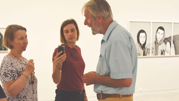 Foto: Drei Menschen unterhalten sich im Ausstellungsraum. Eine Person hält ein Aufnahmegerät, eine andere einen Blindenlangstock.