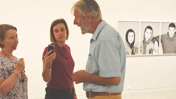 Foto: Drei Menschen unterhalten sich im Ausstellungsraum. Eine Person hält ein Aufnahmegerät, eine andere einen Blindenlangstock.