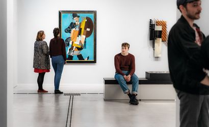 Foto: Mehrere Besucher*innen stehen und sitzen im Ausstellungsraum und betrachten Kunstwerke.