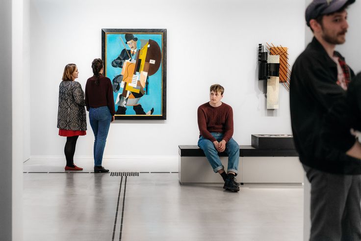 Foto: Mehrere Besucher*innen stehen und sitzen im Ausstellungsraum und betrachten Kunstwerke.