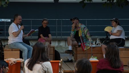 Foto: Blick auf fünf Personen auf Holzbänken. Eine Person blickt auf ein Buch und hält ein Mikrofon in der Hand.