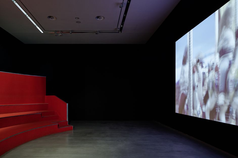 Foto: Eine halbrunde Tribüne mit drei Stufen als Sitzfläche gegenüber einer raumhohen Filmprojektion in einem schwarzen Raum.