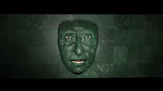 Filmstill: Blick auf eine dunkelgrün-marmoriert geflieste Wand, aus der ein Gesicht mit leicht geöffnetem Mund hervortritt.