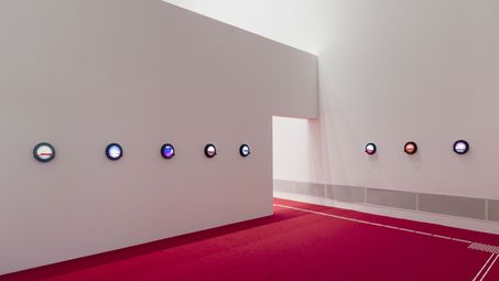 Foto: Blick in den Ausstellungsraum mit einer Reihe Bullaugen mit eingebauten Leuchtkästen an der Wand und leuchtend rotem Teppich.