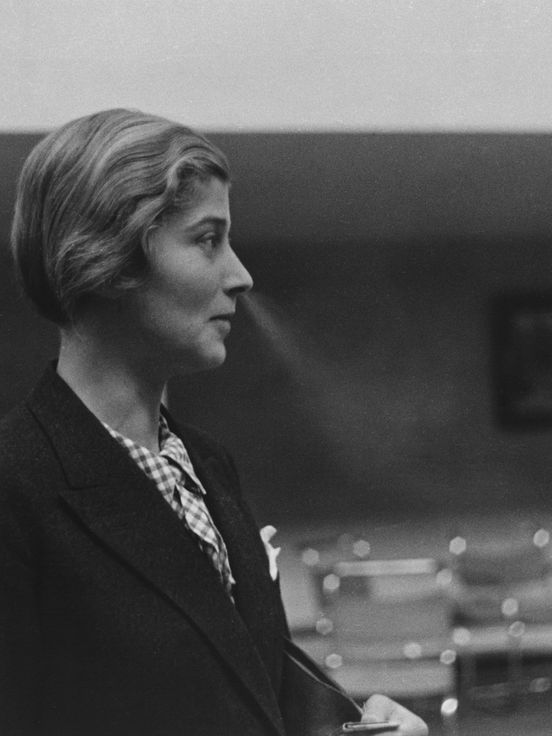 Black and white photograph: side view of a female figure wearing a blazer and checked blouse, exhaling smoke from her nose. The background shows an interior with chairs and a framed picture on the wall.