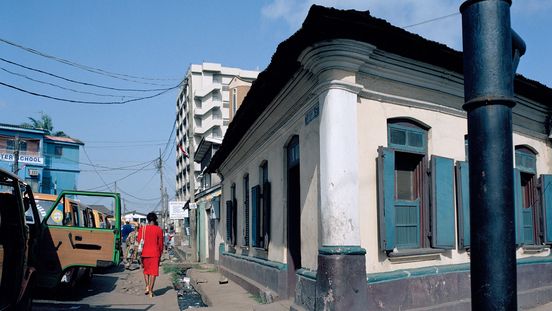 Quadratische Fotografie: Blick auf die vordere Ecke eines hellgelben Gebäudes mit grünen Fensterläden. Rechts daneben läuft eine Person mit rotem Kleid eine schmale Straße entlang.