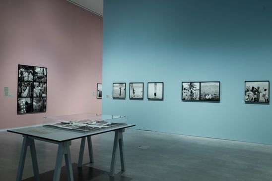 Ausstellungsansicht „Akinbode Akinbiyi“: In einer gläsernen Auslage in der Mitte des Ausstellungsraums liegen Fotobücher. Im Hintergrund sind verschiedene Schwarz-Weiß-Fotografien an den pastel-rosa und pastel-blauen Ausstellungswänden zu sehen.