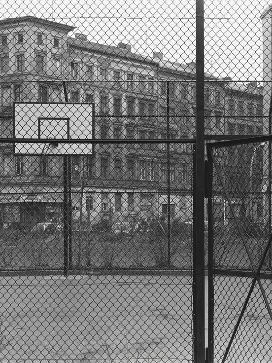 Black-and-white photograph: View of a fenced-in basketball court with the entrance gate open.