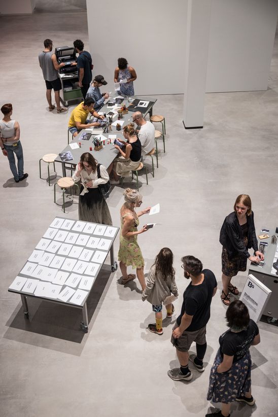 Foto: Einige Zeichen-Workshop-Teilnehmende stehen oder sitzen um zwei Tische und einen Drucker herum.