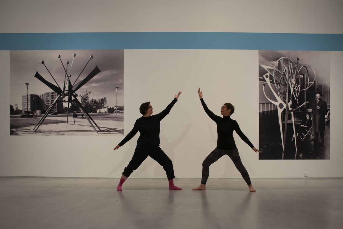 Foto: Zwei Personen in Yoga-Kleidung stehen in Pose vor zwei schwarz-weiß Fototapeten in der Ausstellung „Hans Uhlmann“.