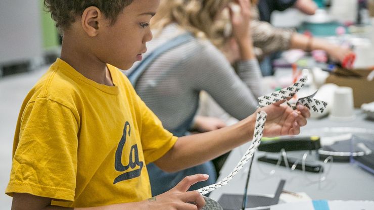 Photo: A child is standing at a table with many craft materials. He is tearing strips off a tape roll.