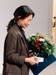 Foto: Jeewi Lee nach der Preisverleihung mit Urkunde, und Blumenstrauß.