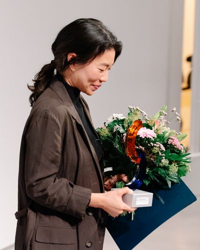 Foto: Jeewi Lee nach der Preisverleihung mit Urkunde, und Blumenstrauß.