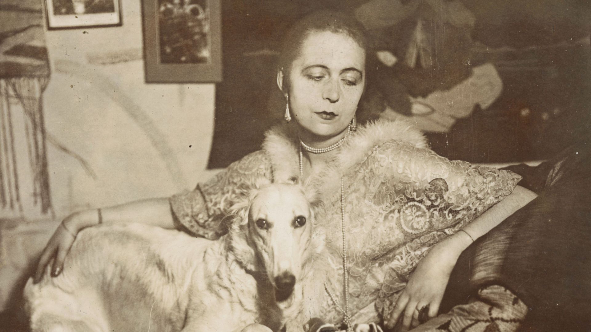 Self-portrait of the artist Marta-Astfalck-Vietz as a black and white photograph. She is sitting on a sofa in a shiny dress with fur trim. A dog with light-coloured fur sits on her lap. Paintings and decorative elements can be seen in the background.