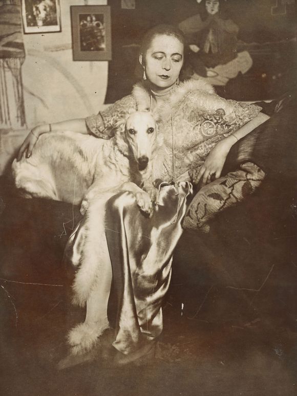 Self-portrait of the artist Marta-Astfalck-Vietz as a black and white photograph. She is sitting on a sofa in a shiny dress with fur trim. A dog with light-coloured fur sits on her lap. Paintings and decorative elements can be seen in the background.