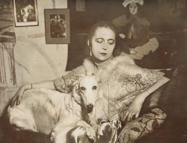 Marta Astfalck-Vietz, Ohne Titel (Selbstporträt), um 1927 Selbstportrait der Künstlerin Marta-Astfalck-Vietz als Sepia-Fotografie. Sie sitzt in einem glänzenden Kleid mit Pelzbesatz auf einem Sofa. Auf ihrem Schoß sitzt ein Hund mit hellem Fell. Im Hintergrund sind Gemälde und dekorative Elemente zu sehen.