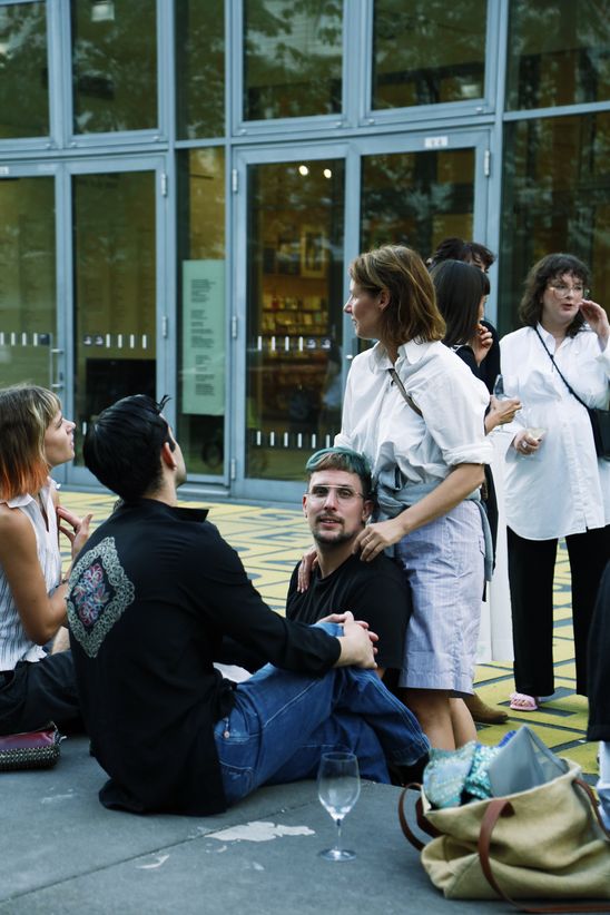 Eine Gruppe von Menschen sitzt draußen, eine stehende Person hat ihre Hände auf eine der sitzenden Personen aufgelegt und blickt zur Seite. 