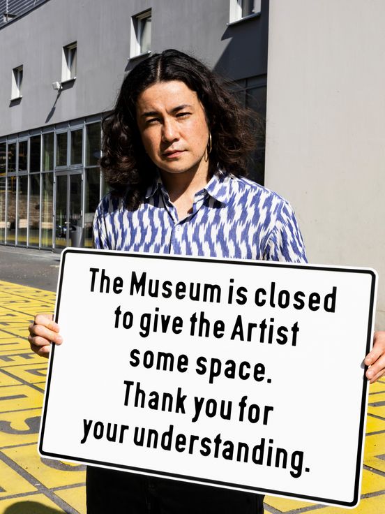 Eine Person steht vor dem Eingang eines Museums und hält ein Schild mit der Aufschrift „The Museum is closed to give the Artist some space. Thank you for your understanding.“ Der Boden ist gelb mit schwarzen Buchstaben bedeckt, im Hintergrund ist eine silberne Skulptur und die Glasfassade des Gebäudes zu sehen.
