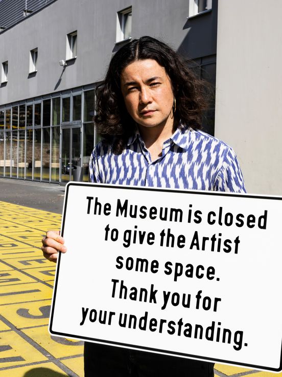 A person stands in front of a museum entrance holding a sign that reads “The Museum is closed to give the Artist some space. Thank you for your understanding.” The ground is covered with yellow tiles featuring black letters, and a silver sculpture and glass façade of the building are visible in the background.