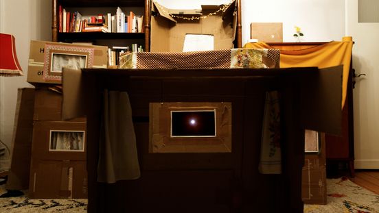 Film still: View of a miniature cinema screen made from cardboard with small curtains and a screen in the shape of a postcard. The background shows a bookshelf and other “cinema boxes”.