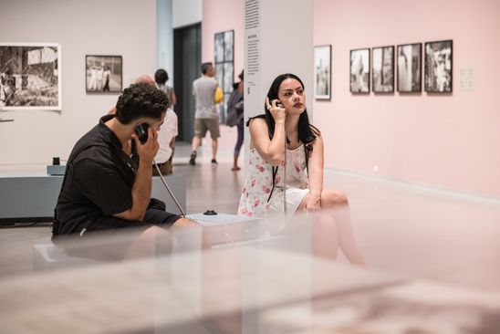 Foto: Zwei Personen sitzen an einer Hörstation im Ausstellungsraum und halten jeweils einen Lautsprecher an ihr Ohr.