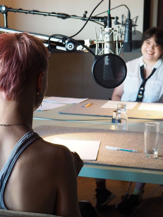 Foto: Zwei junge Menschen sitzen sich in einem Aufnahmestudio gegenüber an zwei Tischen. Vor ihren Köpfen sind Mirkofone installiert. 