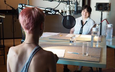 Foto: Zwei junge Menschen sitzen sich in einem Aufnahmestudio gegenüber an zwei Tischen. Vor ihren Köpfen sind Mirkofone installiert. 