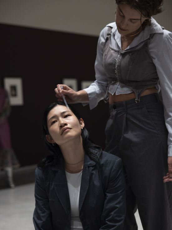 Foto: Deva Schubert steht hinter Chihiro Araki, die auf dem Boden sitzt, und tropft ihr mit einer Pipette Tränen auf die Wange.