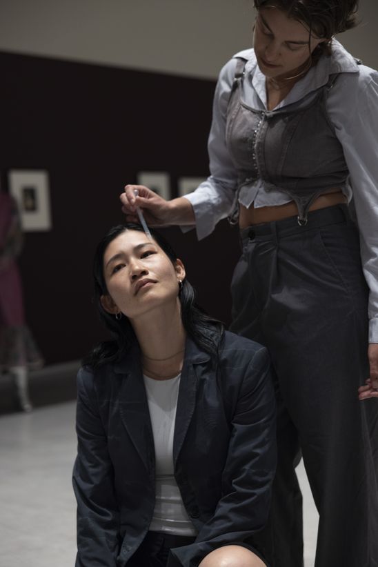 Photo: Deva Schubert stands behind Chihiro Araki, who is sitting on the floor, dripping tears onto her cheek with a pipette.