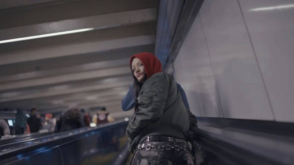 Film still: A person in a bomber jacket and red hoodie is travelling up an escalator. She has her head turned slightly backwards and is looking directly into the camera.