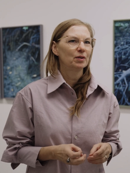 Video still: A person wearing a blouse stands in front of four framed photographs hanging on a wall.