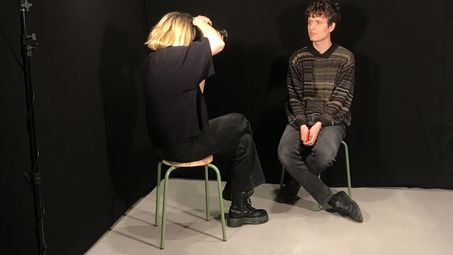 Foto: In einem Raum mit schwarzen Wänden und Scheinwerfern fotografiert eine Person eine andere.