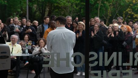 The artist Daniel Hölzl speaks in front of an applauding outdoor audience at the Berlinische Galerie.