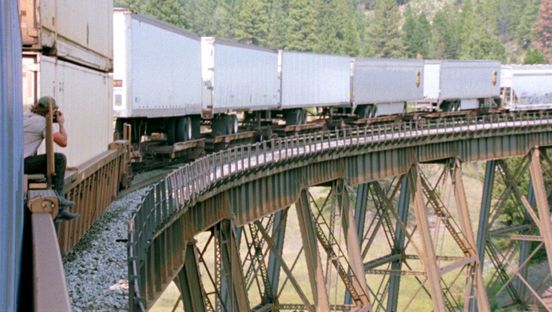 Film still: A person sits on the right of the picture on a goods train making a turn, holding a camera.