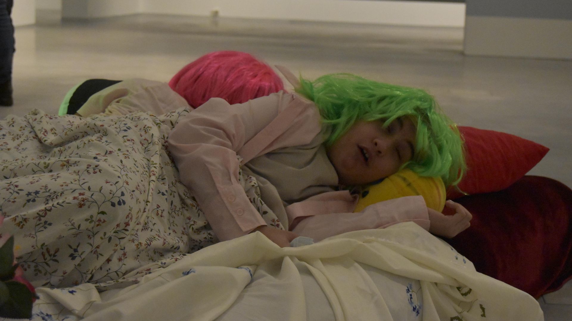 Foto: Kind mit grüner Perrücke hat sich ein Bett im Ausstellungsraum gebaut und gibt vor zu schlafen.. 