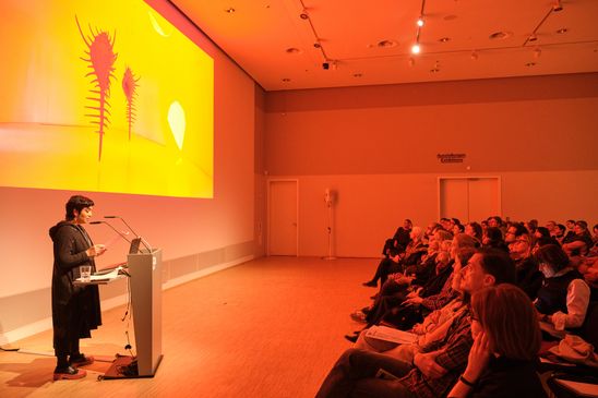 Photo: Monira Al Qadiri stands at a lectern in front of an audience. The room is bathed in warm, orange light from her presentation projected onto the wall.
