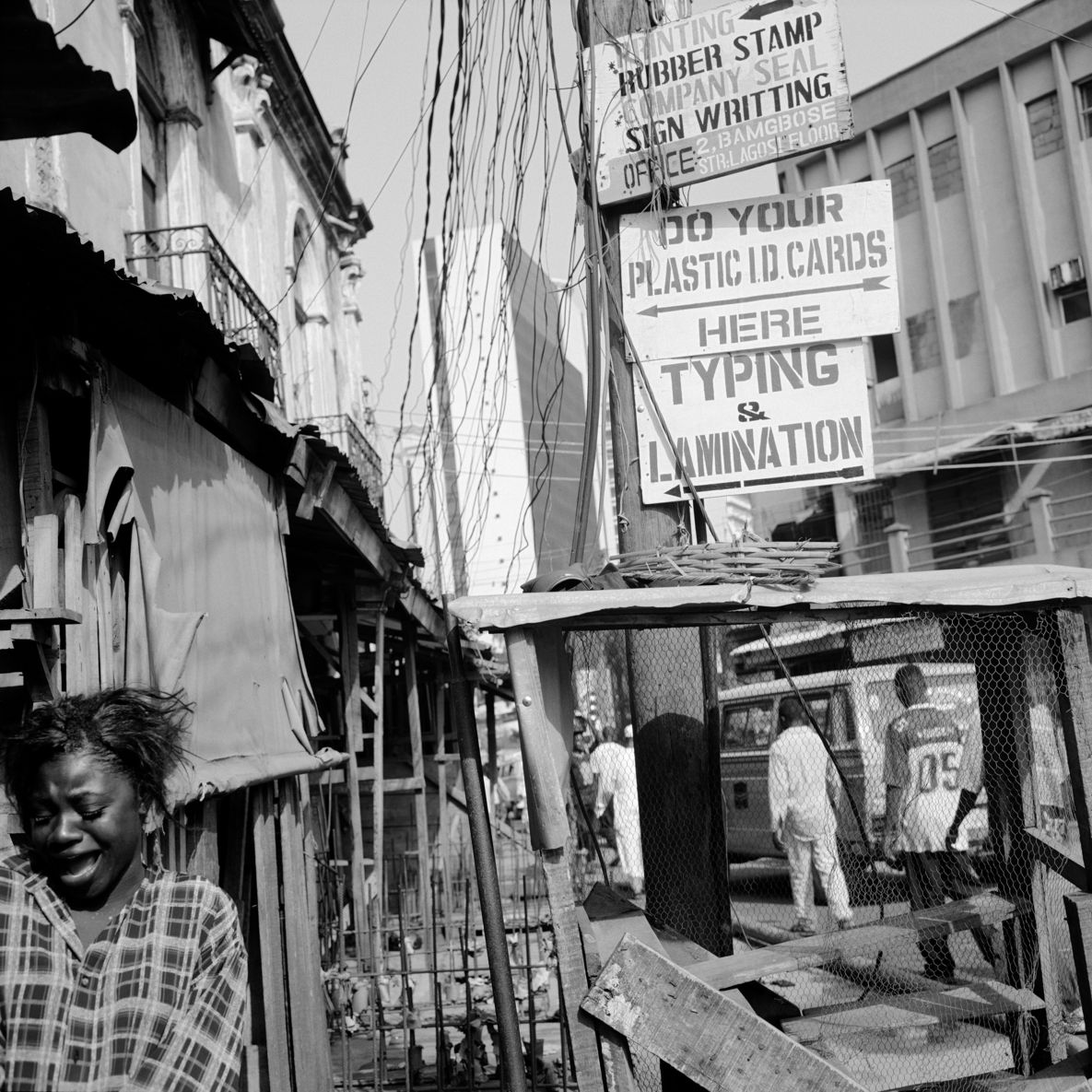 Square black and white photograph: View of various house facades with a street running between them and people walking alongside. An electricity pylon can be seen in the center of the picture, to which billboards with inscriptions such as “Typing & Lamination” are attached. In the front, left-hand part of the photograph, a female figure can be seen with her mouth wide open and her eyes downcast.