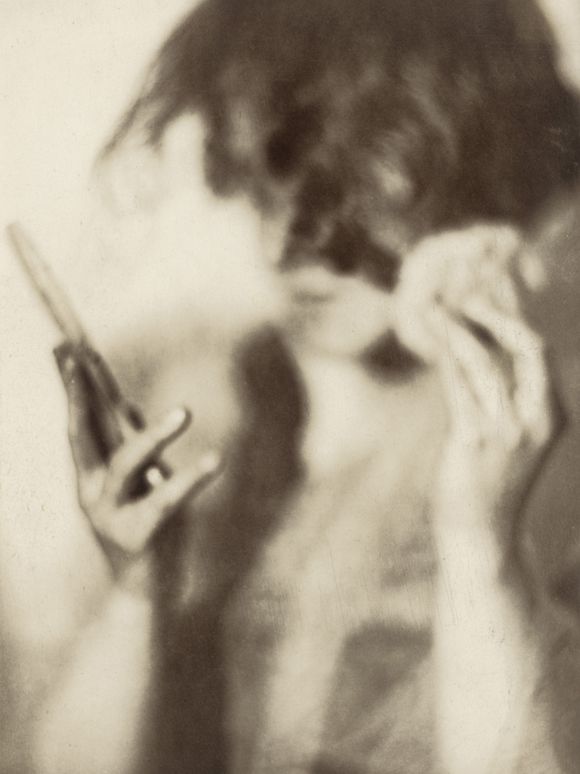 Sepia photograph: A female figure looks sideways at a small mirror, which she holds in her right hand. In her left hand she holds a small powder puff.