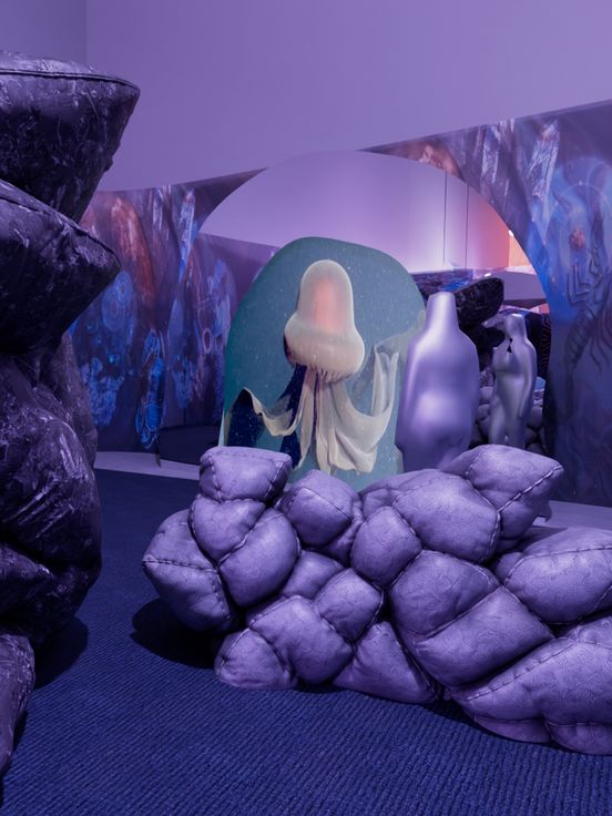 Digitales Rendering einer Installation mit großen, dunklen Kissenstapeln und einer weißen Skulptur in einer lila beleuchteten Umgebung.