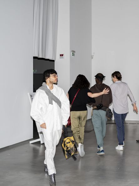 Photo with flash: Front view of a person in white clothing and glittering silver shoes walking out of an exhibition room, while three people walk into the room in the background.