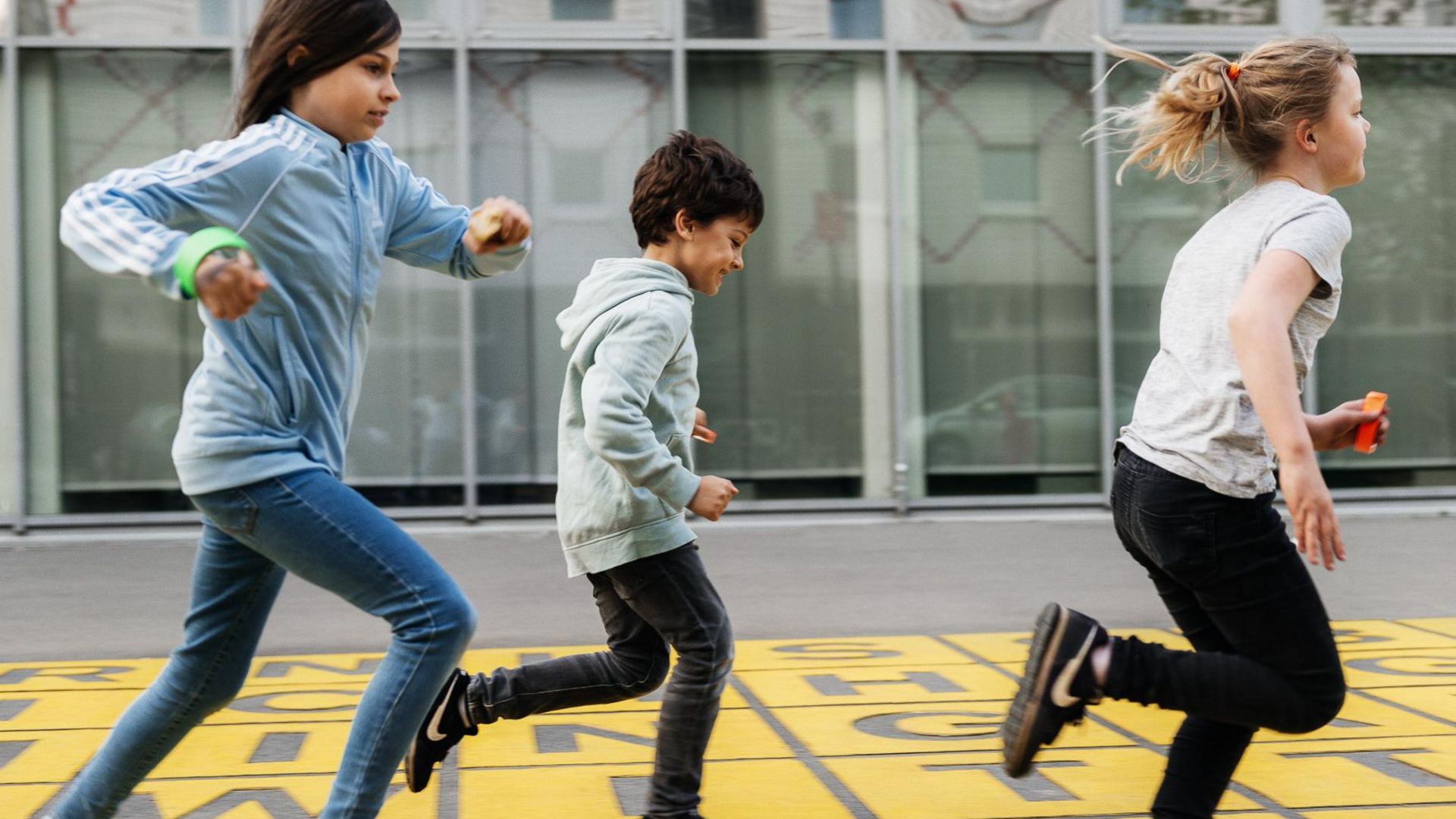 Photo: Three children run across a piece of floor art in front of the museum: grey letters on a yellow background.
