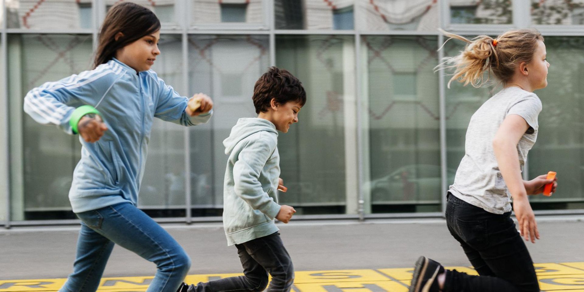 Foto: Drei Kinder rennen über ein Bodenkunstwerk vor dem Museum: Graue Buchstaben auf gelbem Untergrund.