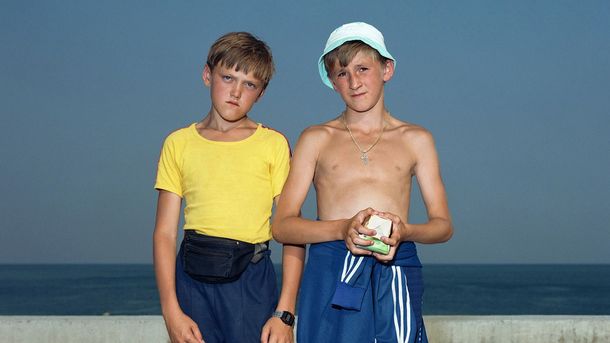 Photo: Two boys stand in front of a low concrete balustrade with the sea in the background. Both are wearing sportswear and looking directly into the camera.