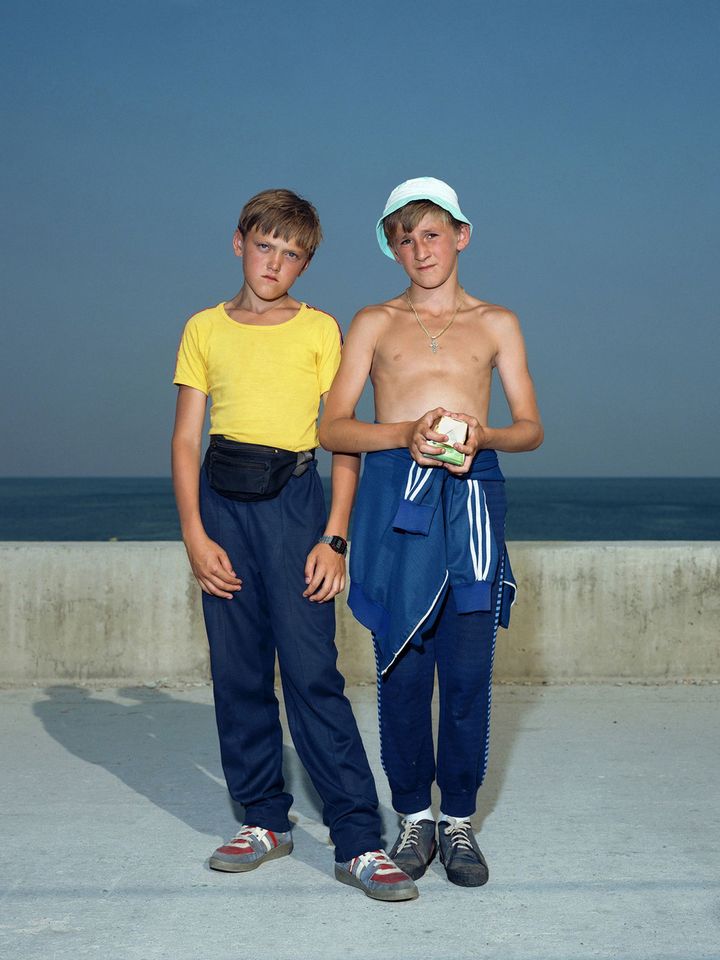 Photo: Two boys stand in front of a low concrete balustrade with the sea in the background. Both are wearing sportswear and looking directly into the camera.