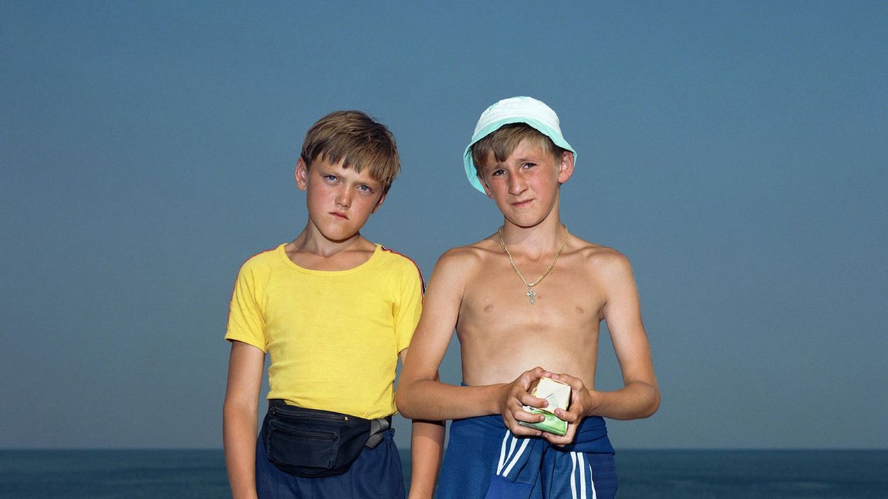 Photo: Two boys stand in front of a low concrete balustrade with the sea in the background. Both are wearing sportswear and looking directly into the camera.