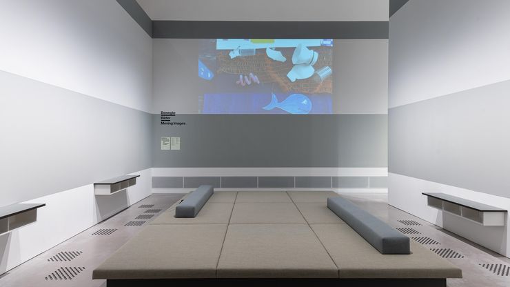 Photo: In the centre of a room is a large upholstered seating and reclining area. Sideboards on the walls to the right and left. An image is projected onto the centre wall.