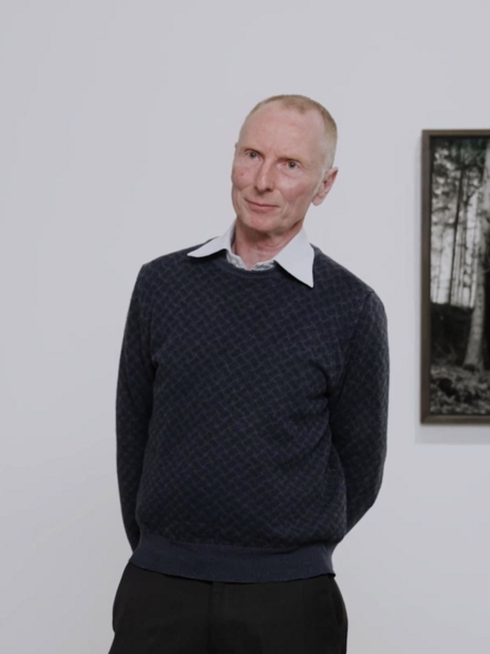 Video still: A person in a dark jumper and shirt collar stands in front of three framed photographs.