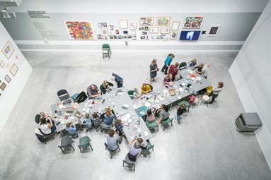 Foto: Blick von oben: Viele Kinder und Erwachsene stehen und sitzen an einem Tisch mit vielen Bastelmaterialien. Sie basteln und unterhalten sich. Viele gemalte Bilder und Zeichnungen hängen an den Wänden.